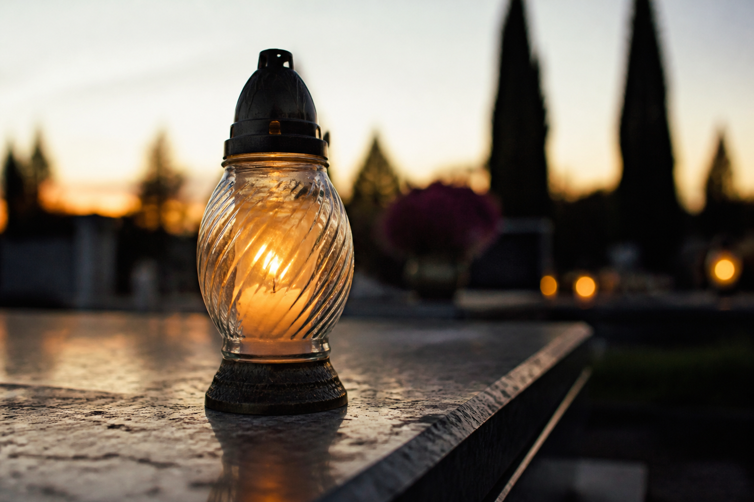 Palący się szklany znicz stoi na kamiennej płycie nagrobnej o zmierzchu, tło cmentarza jest nieostre.